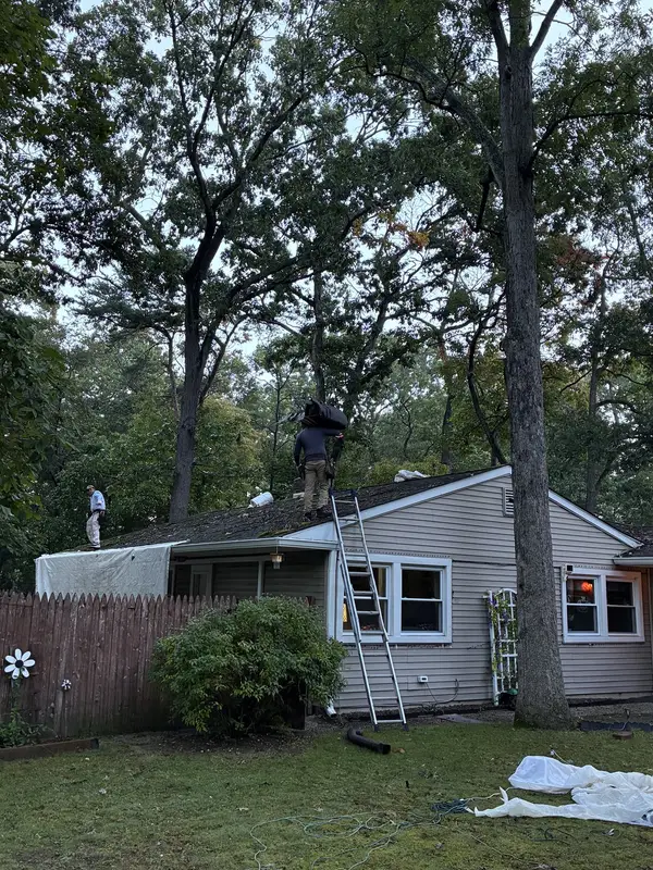 Roofers working on the roof of a tan ranch home, with tarps on the lawn and a ladder at the eave