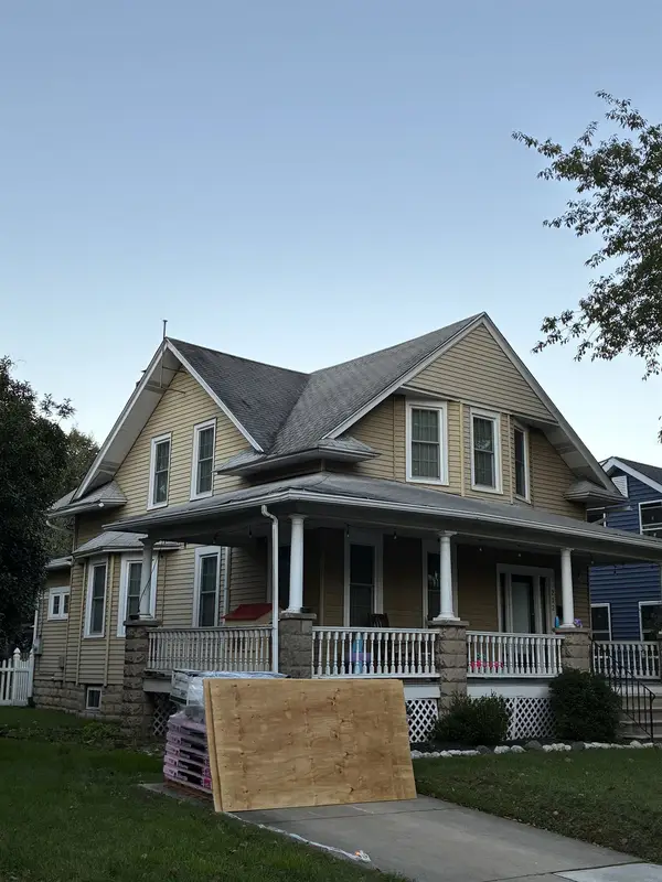 Yellow two-story home with a wraparound porch, with shingle bundles and plywood delivered to the driveway